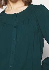 Chemisier bleu sarcelle à col arrondi, avec des détails plissés aux épaules et des manches longues. Le tissu semble léger et lisse.