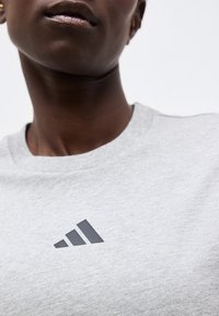 Κοντινό πλάνο ενός ατόμου που φοράει ανοιχτό γκρι πουκάμισο με μαύρο λογότυπο Adidas στο στήθος.