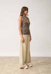 Brązowy bezrękawnik z głębokim dekoltem w kształcie V, z guzikami z przodu i kieszeniami; w połączeniu z kremowymi, szerokimi, prążkowanymi spodniami i jasnymi trampkami.