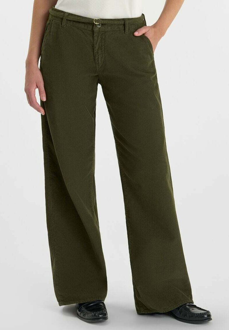 Persona que lleva puestos pantalones de pana verde oliva de pierna ancha con un cinturón delgado, una blusa blanca y zapatos negros, de pie con una mano en el bolsillo.