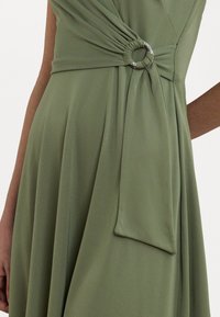 Vestido verde com um design torcido e um acento em anel prateado circular. Tecido suave com pregas drapeadas na saia.