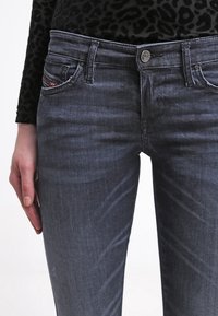 Jeans de mezclilla oscura con un diseño ajustado, costuras visibles, un cierre de botón en la parte frontal y cuatro bolsillos. La textura parece suave con un ligero brillo.