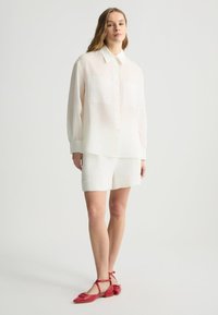 Camicia bianca a maniche lunghe con bottoni, dotata di due tasche sul petto, abbinata a pantaloncini bianchi e scarpe rosse basse con cinturini alla caviglia.