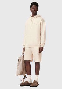 Completo con felpa beige e pantaloni corti, dotato di tasca frontale e logo coordinato. Indossato con calze beige e scarpe scivolate marroni, mentre tiene uno zaino beige chiaro.