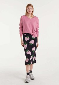 Suéter rosa de manga larga combinado con una falda negra a la altura de la rodilla que presenta un patrón de fresas en rosa y verde. Lleva zapatillas grises.