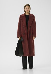 Cappotto doppiopetto in misto lana rosso con un ampio colletto e superficie testurizzata. Abbinato a pantaloni neri a gamba larga e una borsa nera.