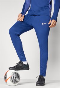 Modré športové nohavice vyrobené z hladkého materiálu, s oranžovým logom Nike, v kombinácii s čiernymi topánkami na bielej vzorovanej futbalovej lopte.