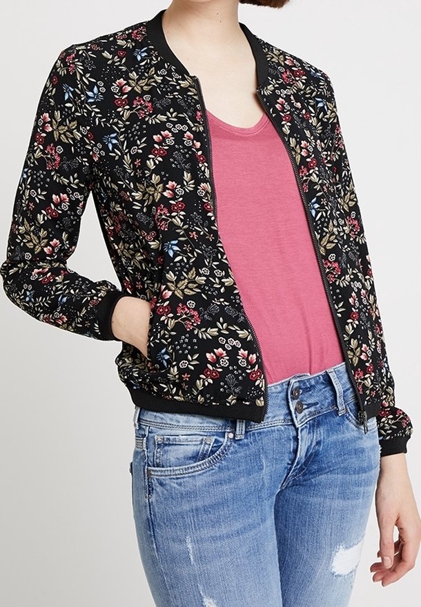 Blouson aviateur noir avec imprimé floral en rouge, rose et vert. Poignets et ourlet côtelés, fermeture éclair, associé à un haut rose et un jean bleu clair.