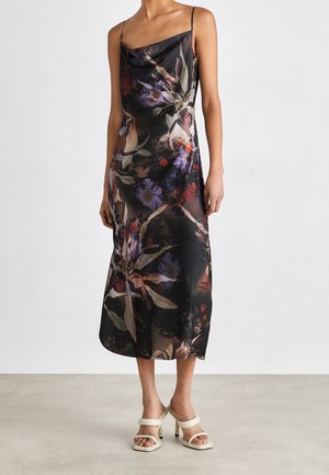 Femme portant une robe midi noire sans manches avec un imprimé floral violet et rouge, et des sandales blanches à talons hauts, debout sur un sol uni.