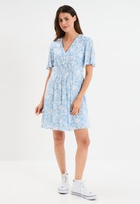 Hellblaues Kleid mit floralem Muster, kurzen Ärmeln, V-Ausschnitt, smockierter Taille und knielangem Saum. Kombiniert mit weißen High-Top-Sneakern.