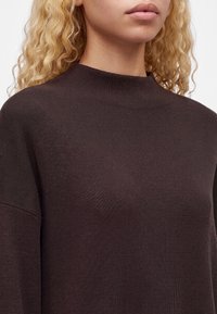 Personne blonde portant un pull en maille côtelée marron foncé à col montant, montrée des épaules à la bouche.