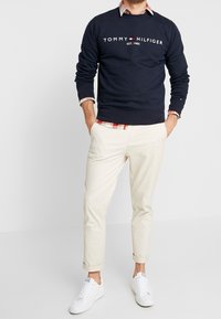 Felpa blu navy con testo bianco, logo a toppa e polsini a coste. Abbinate a pantaloni crema e sneakers bianche, che mostrano un motivo a scacchi stratificato.