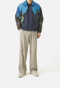 Giubbotto bomber colorato con un design floreale, t-shirt blu navy, pantaloni a gamba larga beige e sneaker bianche. Il giubbotto ha polsini e colletto elasticizzati.