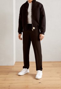 Svart bomberjacka med ribbade muddar, dragkedja framtill, i kombination med svarta byxor och vita sneakers på trägolv. Texturerade tygdetaljer synliga.