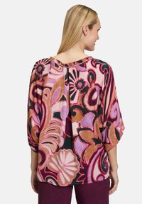 Farverig bluse med blomster- og abstrakte mønstre i pink, rød og grøn, med løse ærmer og detalje med en plissering bagpå.
