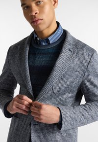 Blazer a motivo houndstooth grigio e blu con revers a punta, chiusura a un bottone e taschino sul petto, indossato sopra un maglione blu navy testurizzato.