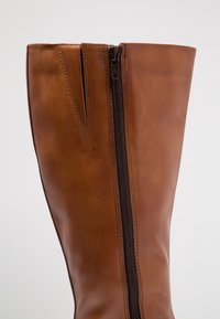 Botte en cuir marron jusqu'au genou avec une texture lisse, dotée d'une fermeture éclair sur le côté et de détails de surpiqûres contrastants. Design structuré mais flexible.