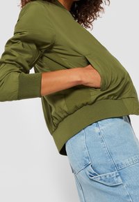 Veste bomber vert olive avec une texture lisse, des poignets côtelés et une poche latérale ; associée à un jean taille haute bleu clair.