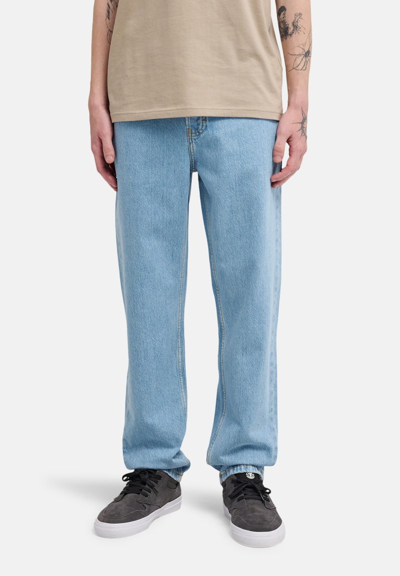 Persona vistiendo jeans holgados azul claro, camiseta beige y zapatillas negras con suelas blancas, de pie contra un fondo blanco.