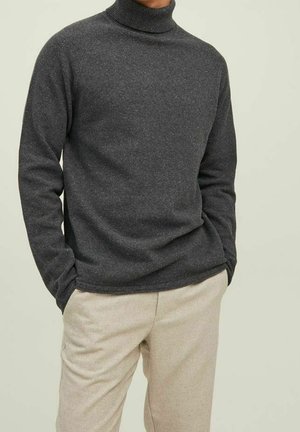 Homme portant un pull à col roulé gris foncé avec les mains dans les poches et un pantalon beige clair sur un fond uni.