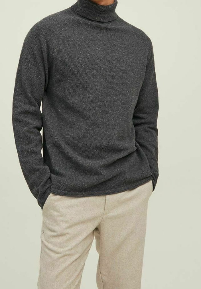 Homme portant un pull à col roulé gris foncé avec les mains dans les poches et un pantalon beige clair sur un fond uni.