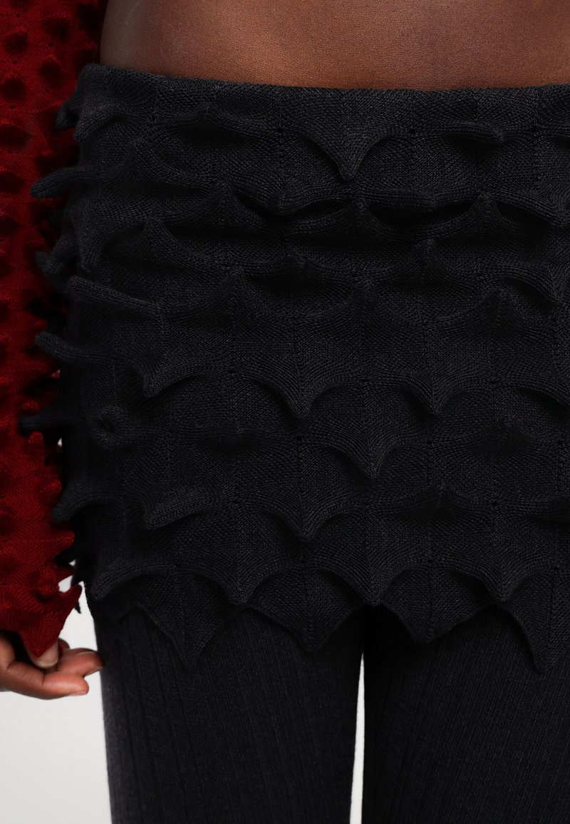 Schwarzer Strickrock mit texturiertem, gewelltem Design und geschichteten Akzenten. Kombiniert mit gerippten schwarzen Leggings und einem roten Oberteil mit Puffärmeln.