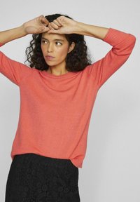 Korallfarget strikkegenser med scoop-hals og trekvartslange ermer, kombinert med et svart blondeskjørt, mot en nøytral bakgrunn.