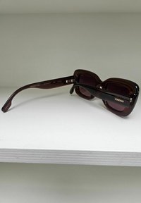 Rectangulaire bruine zonnebril met een glanzende afwerking. De pootjes hebben een contrasterende zwarte kleur met "KOMONO"-branding in goud.