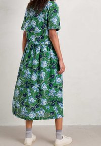 Robe florale verte à manches courtes, ornée de motifs de fleurs bleues et blanches, avec une taille froncée et une longueur mi-mollet. Associée à des baskets blanches.
