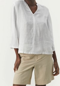 Personne portant un haut en lin blanc à col en V, à manches longues, avec un short beige jusqu'aux genoux devant un fond uni.