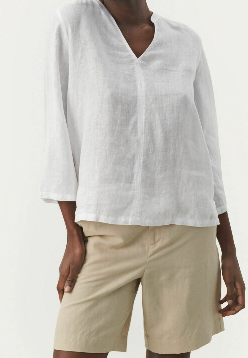 Personne portant un haut en lin blanc à col en V, à manches longues, avec un short beige jusqu'aux genoux devant un fond uni.