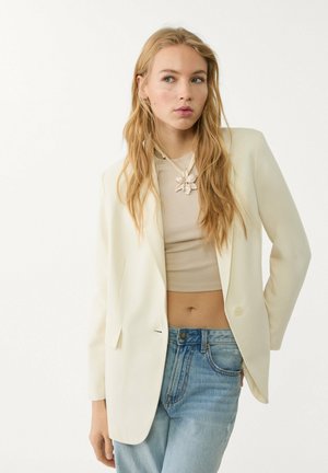 Blazer u boji kreme, krojen na telo, sa jednim dugmetom, uparen sa bež crop topom i svijetloplavim farmerkama visokog struka. Duga, talasasta kosa.