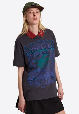 Joven mujer con una camiseta gráfica oscura con diseño azul y verde, cuello rojo, falda negra y gorra Vans verde oliva.