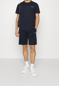 Mężczyzna ubrany w granatowy t-shirt Calvin Klein oraz szorty, w białych skarpetkach, białych sportowych butach i czarnej torbie na ramię, stojący na tle białym.