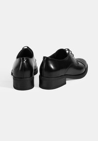 Chaussures en cuir noir avec une forme arrondie, finition brillante, petit talon empilé et perforations décoratives sur la zone des orteils.