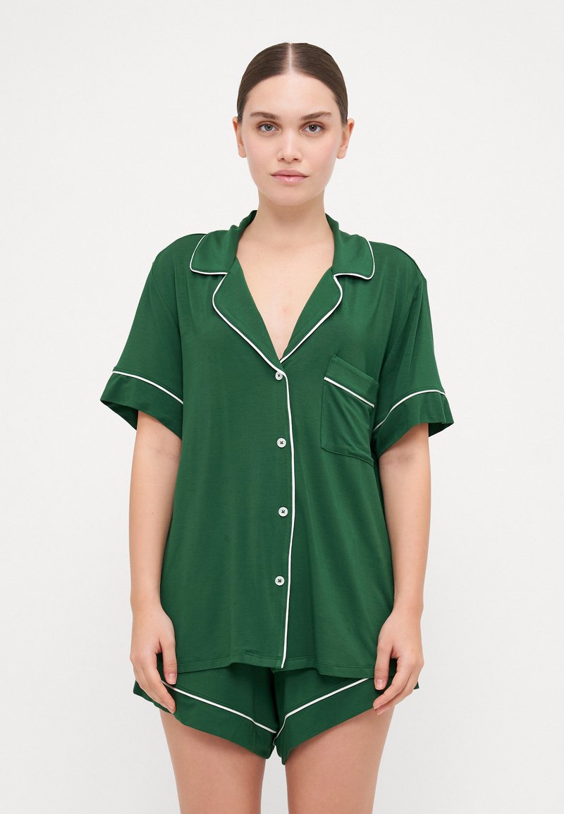 Conjunto de pijama de manga curta verde feito de tecido suave. Apresenta acabamentos brancos, um bolso no peito e uma frente com botões com um corte descontraído.