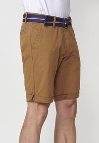 Homme portant un short marron juste au-dessus du genou avec des ourlets retroussés et une ceinture bleu marine avec des rayures rouges et blanches, se tenant devant un fond uni.