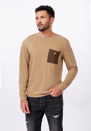 Beige long-sleeve shirt with a brown chest pocket featuring a yellow logo. The fabric appears soft, with ribbed cuffs and a round neckline.