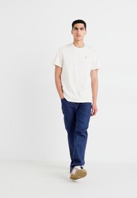 Hvit bomull t-skjorte med en liten logo, kombinert med blå denimjeans og lyse sneakers. Modell som står mot en ensfarget bakgrunn.