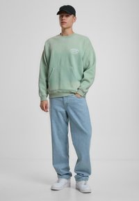 Lys grønn bomull sweatshirt med frontlomme, kombinert med lyseblå avslappede jeans og hvite sneakers. Svart caps fullfører antrekket.