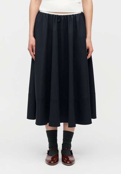 Γυναίκα που φοράει μαύρη φούστα μέχρι το γόνατο, σκουρόχρωμες γκρι κάλτσες και σκοτεινά κόκκινα παπούτσια Mary Jane, στέκεται μπροστά σε ένα απλό λευκό φόντο.