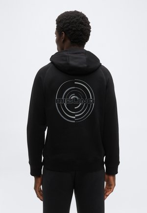 Άνδρας που φοράει μαύρο φούτερ με κουκούλα με κυκλικό σχέδιο και κείμενο "Mizuno" στην πλάτη, στέκεται μπροστά σε απλό ανοιχτό φόντο.