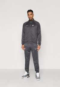 Mand står iført en mørkegrå Chicago Bulls Nike træningsdragt og hvide sportsko foran en lys grå baggrund.