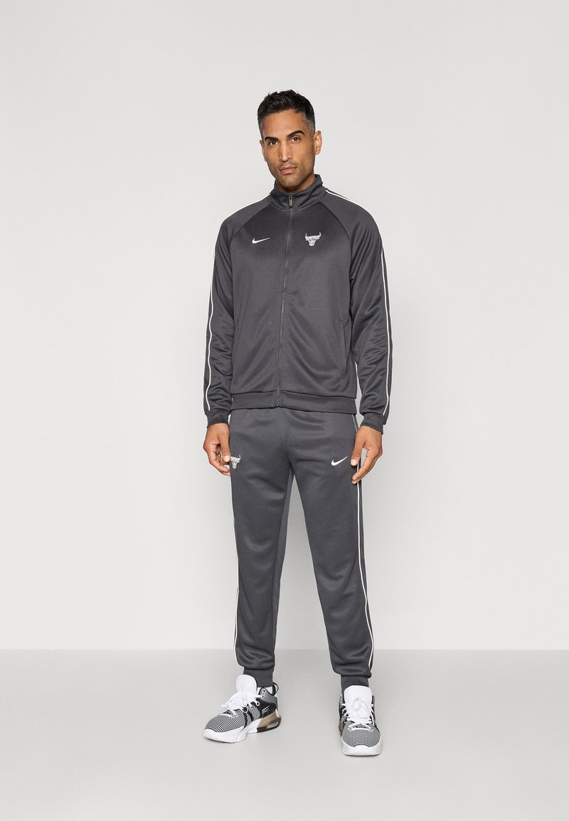 Mand står iført en mørkegrå Chicago Bulls Nike træningsdragt og hvide sportsko foran en lys grå baggrund.