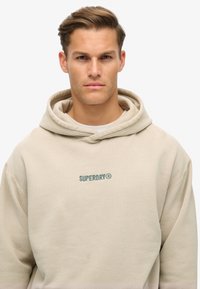 Sudadera beige con capucha ajustable, que presenta un logo bordado en verde "SUPERDRY®" en el pecho. Tejido suave con un corte relajado.