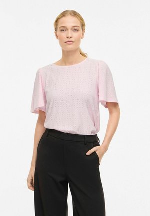 Femme aux cheveux blonds attachés en queue de cheval, portant un chemisier léger rose à manches courtes et un pantalon noir taille haute, debout avec une main dans la poche.