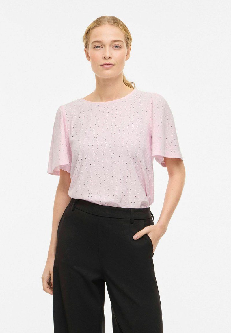 Žena s blond vlasmi v copu, oblečená v svetloružovej krátkorysej blúzke a čiernych vysokoprstových nohaviciach, stojaca s jednou rukou v vrecku.