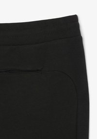 Un pantalon de jogging noir présente une texture lisse, une taille élastique et une poche zippée dissimulée sur le côté pour un rangement sécurisé.