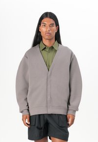 Grå oversized tröja med V-hals och raglanärmar, lagd över en olivgrön skjorta med knappar. Svarta shorts fulländar looken.