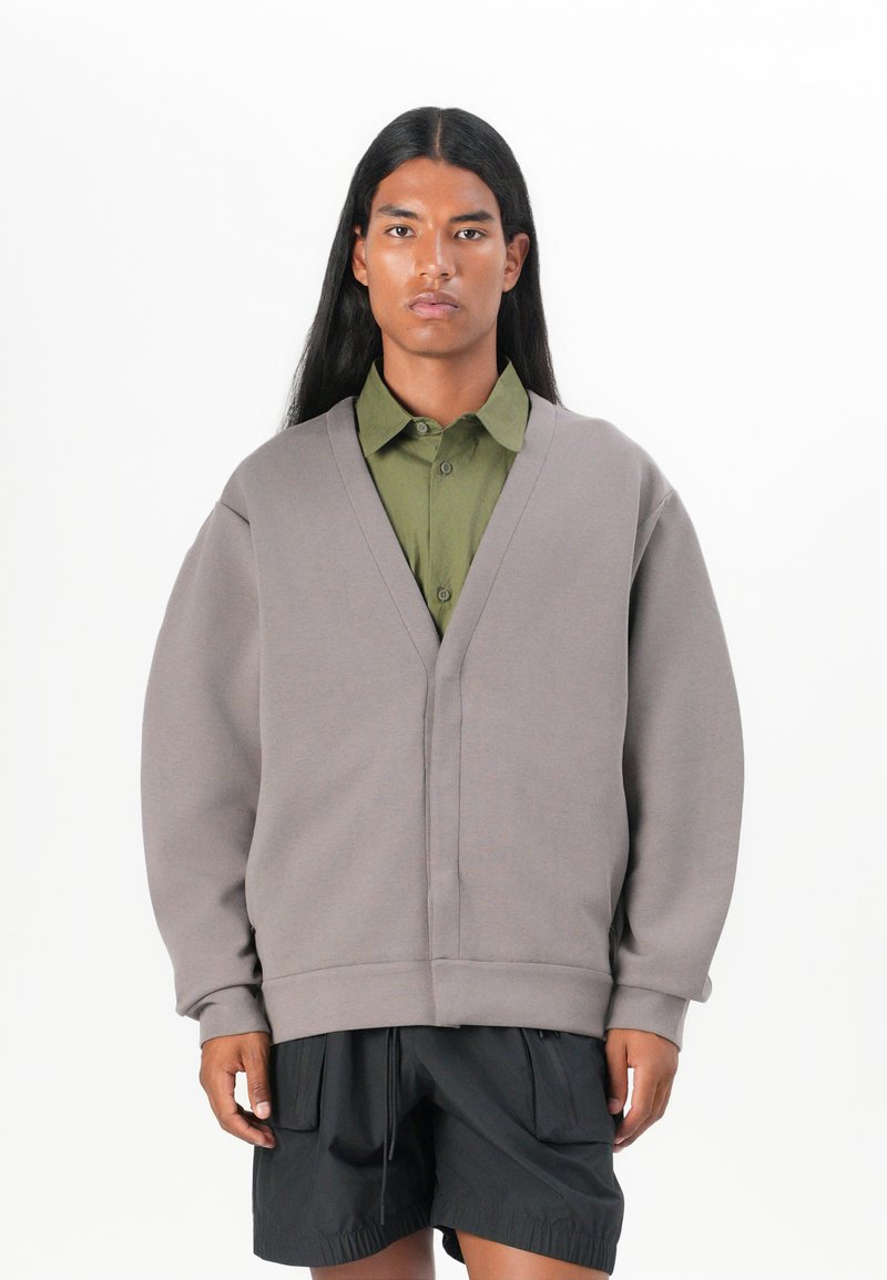 Grå oversized tröja med V-hals och raglanärmar, lagd över en olivgrön skjorta med knappar. Svarta shorts fulländar looken.